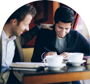 Image of two men at a coffee shop, one has his hand on the other's shoulder in support while the other one is visibly writing something on some notebooks in front of him